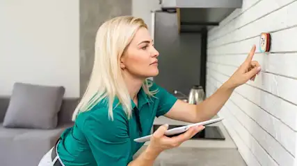 Vrouw met tablet bedient thermostaat op witte bakstenen muur in keuken. Koelkast en waterkoker zichtbaar.