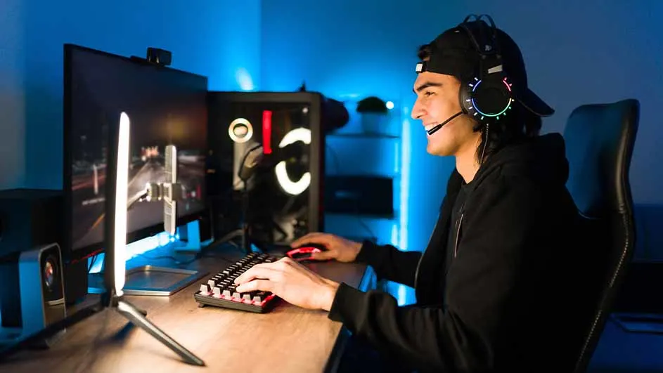 Een persoon met een headset zit achter een bureau met een toetsenbord, muis en computerschermen in een blauwe omgeving.