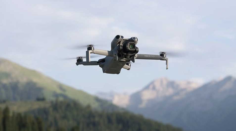 A grey drone hovers mid-air, set against a backdrop of green mountains and blue skies, ready to capture photos. 