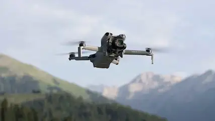 A grey drone hovers mid-air, set against a backdrop of green mountains and blue skies, ready to capture photos.