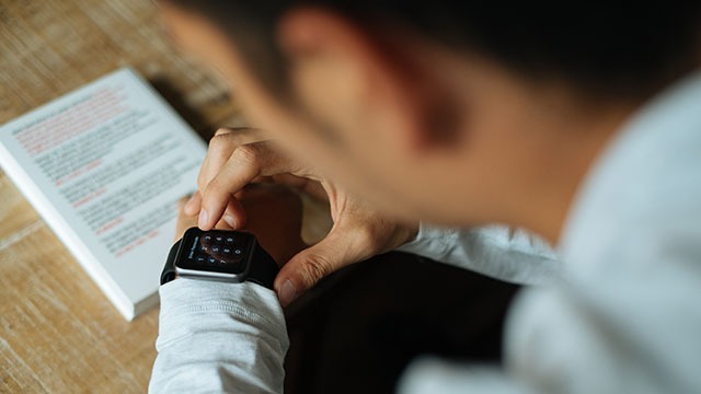 Person in a white shirt uses a black smartwatch on a wooden surface next to a white book.