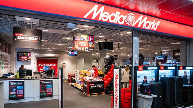 Ingang van een Media Markt winkel met rode reclameborden en "Black Friday" promoties. Binnen zijn schappen en elektronica zichtbaar.