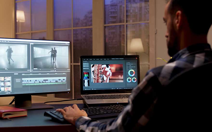 Een man bewerkt video’s op een laptop met een tweede scherm in een donkere kamer.