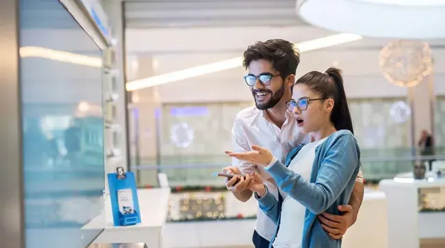 Een man en een vrouw met brillen kijken naar televisies in een winkel, de vrouw wijst met haar hand.