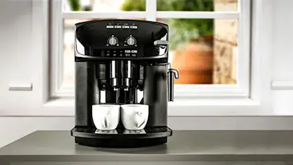 A black coffee maker sits on a counter with two white cups, set against a window showing greenery.