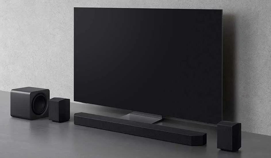 Een grote zwarte flatscreen tv staat op een grijze ondergrond, met een soundbar, subwoofer en twee achterspeakers in strak zwart design ervoor.