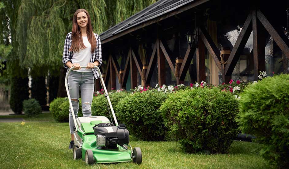 Een vrouw maait het gras met een groene grasmaaier in een tuin met struiken en een houten huis op de achtergrond. Het merk is niet zichtbaar.