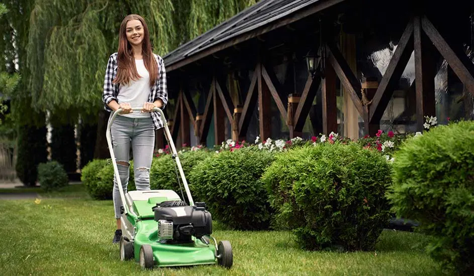 Een vrouw maait het gras met een groene grasmaaier in een tuin met struiken en een houten huis op de achtergrond. Het merk is niet zichtbaar.