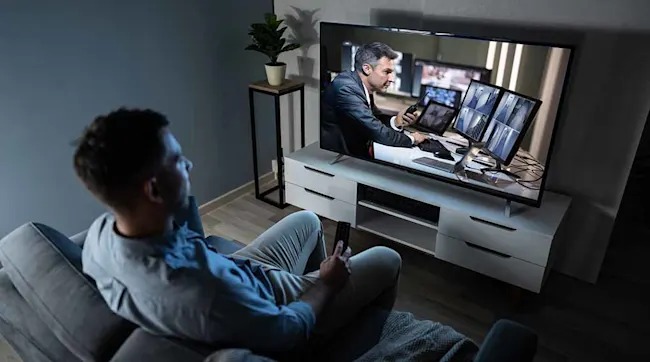 Een man zit op een grijze bank met een afstandsbediening, kijkend naar een tv waarop een man achter een bureau te zien is.