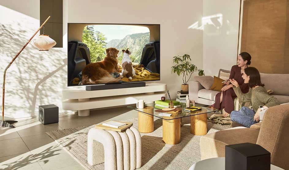 Een moderne woonkamer waar twee vrouwen naar een groot scherm kijken met honden in berglandschap. Houten tafeltjes, beige fauteuils, plant, natuurlijk licht. Gezellige, warme huissfeer met technologie.