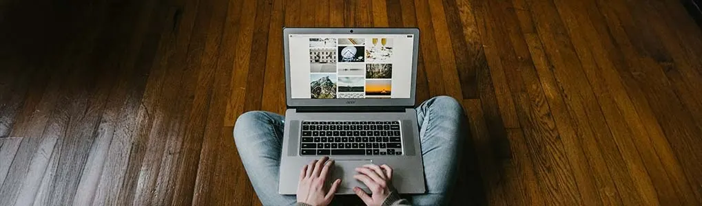 Persoon zit op een houten vloer met een laptop op schoot en typt. Het scherm toont een fotogalerij.