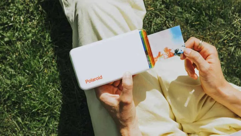Een persoon zit in het gras, met een witte Polaroid printer en een foto in de hand.