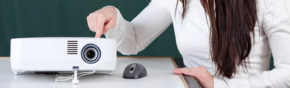 A woman with long brown hair and a white top is sitting at a table, pressing a button on a white projector. No brand name is visible on the device.