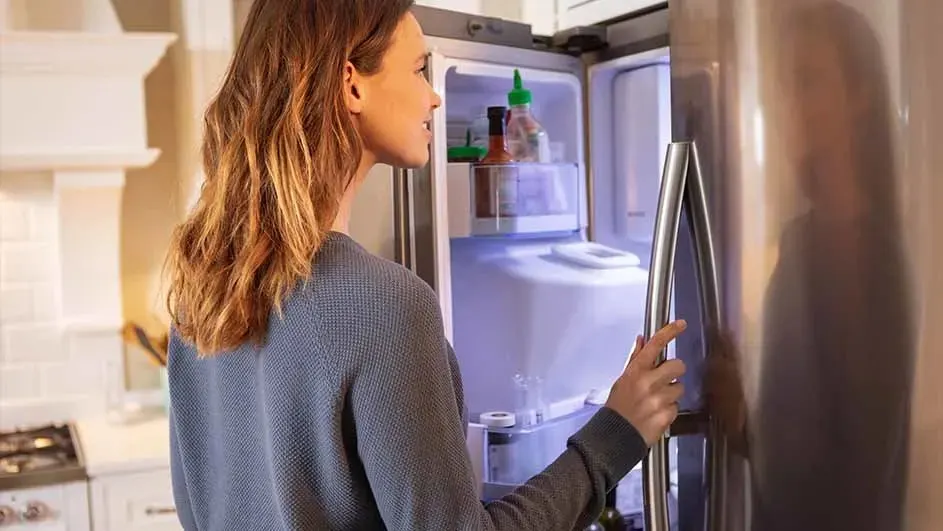 Een vrouw opent een grote, zilveren koelkast in een lichte keuken. In de koelkast staan sauzen en een ijsmaker. Het design is modern.
