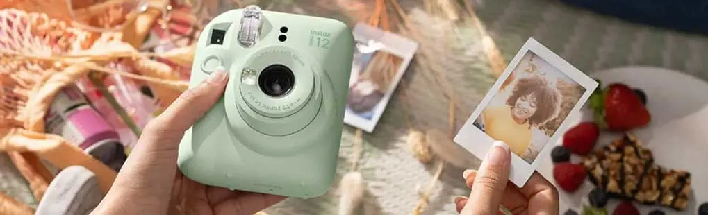 Een hand houdt een Instax-camera, terwijl de andere hand een polaroidfoto vasthoudt. Op de achtergrond fruit en snacks.