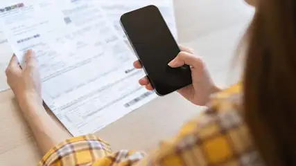 Vrouw houdt document en zwarte smartphone vast. Ze draagt een geel geruit overhemd.