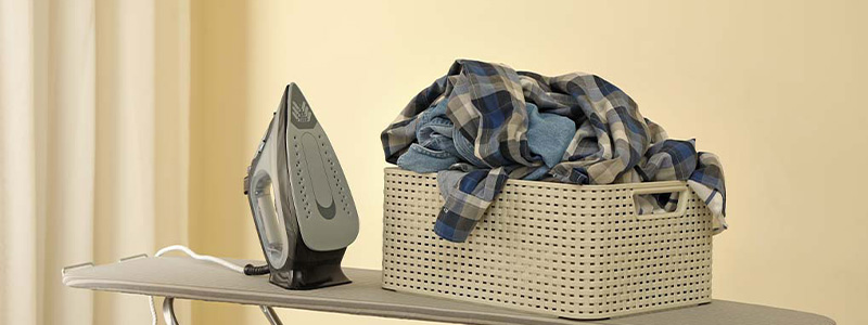 Een strijkijzer naast een mand met blauwe en grijze kleding op een strijkplank tegen een gele achtergrond.