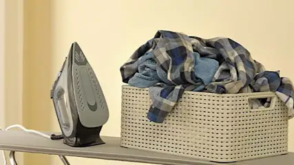Een strijkijzer naast een mand met blauwe en grijze kleding op een strijkplank tegen een gele achtergrond.