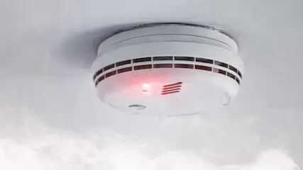 Een rookmelder aan een wit plafond met een rood waarschuwingslampje en rook in de omgeving.