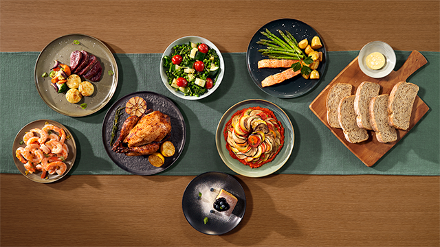 Diverse gerechten, zoals vlees, salade, kip, groenten, brood en garnalen, staan op een blauwgroene loper op een houten tafel.