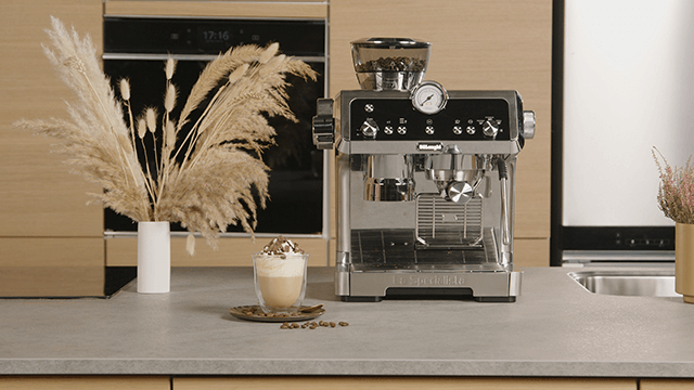 Een zilveren espressomachine staat naast een glas koffie met slagroom op een aanrecht met decoratie.