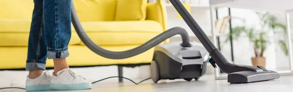 Someone is vacuuming the light floor in a living room with a cylinder vacuum cleaner; a yellow sofa and white table are in the background.