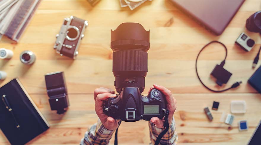 Twee handen houden een zwarte camera vast, omringd door camera-accessoires op een houten tafel.