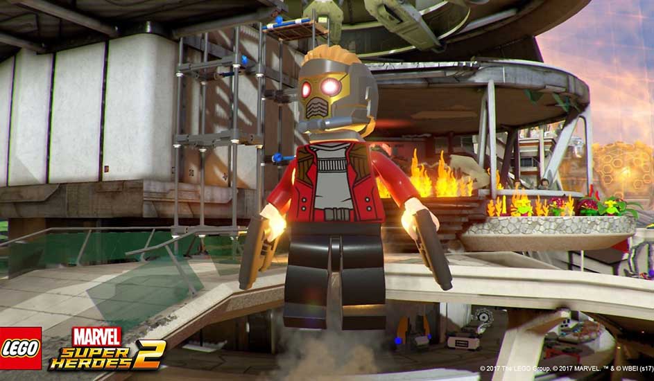 This is a video game. A LEGO superhero floats with blasters in hand above a futuristic building with flames burning in the background.