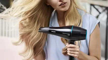 Woman with long blonde hair blow dries her flowing locks using a black Philips hairdryer, creating smooth, shiny movement around her shoulders.