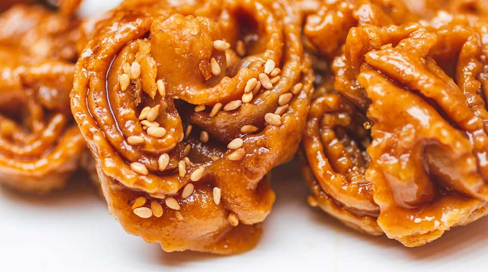 Drie gefrituurde, goudbruine honingkoekjes, versierd met sesamzaadjes, liggen op een witte ondergrond.