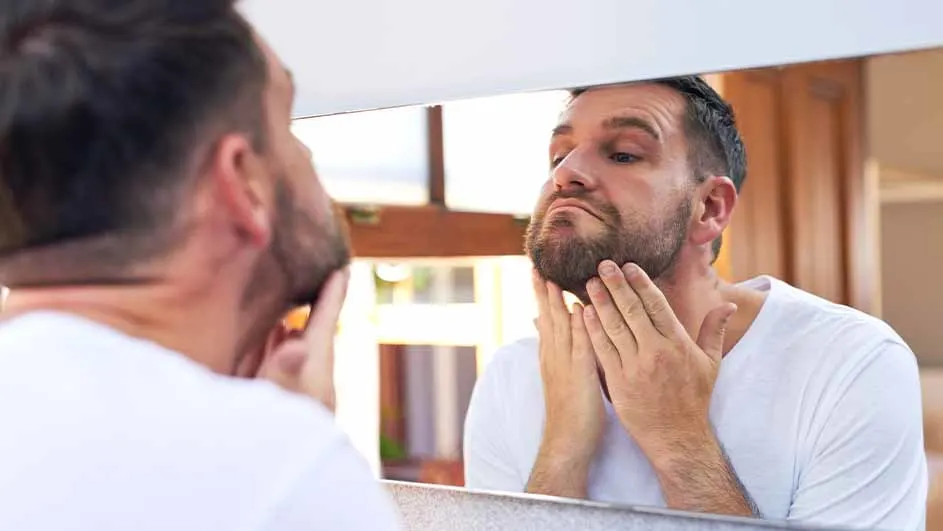 Een man met een baard bekijkt zijn gezicht in een spiegel, terwijl hij zijn kin met beide handen aanraakt.