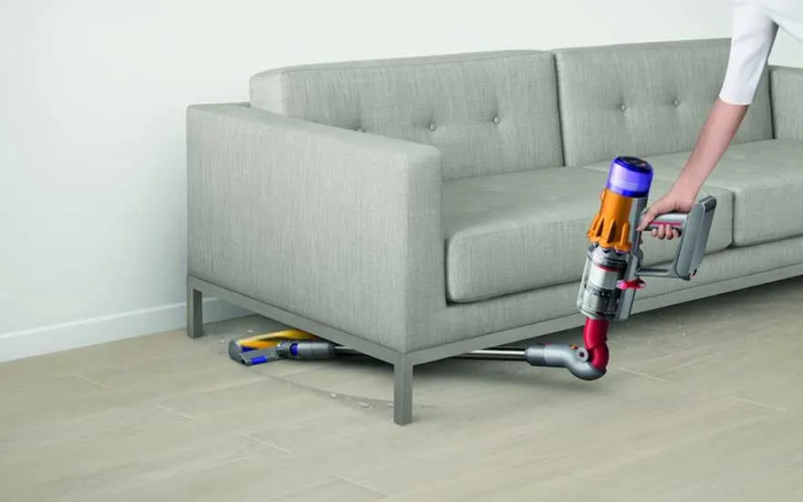Een krachtige steelstofzuiger schuift moeiteloos onder een grijze bank en zuigt stof en kruimels weg op de lichte houten vloer.