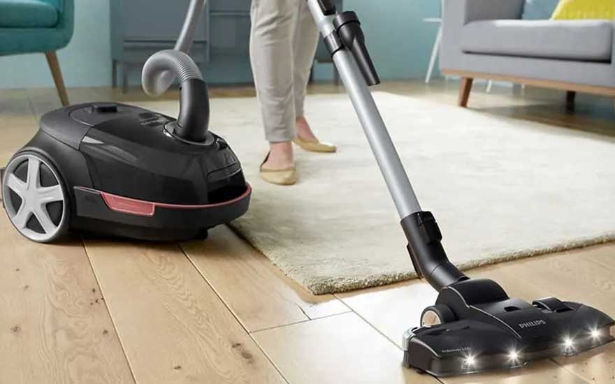 Een zwarte stofzuiger met grote wielen zuigt vuil op een houten vloer, met LED-verlichting voor beter zicht bij het schoonmaken. 