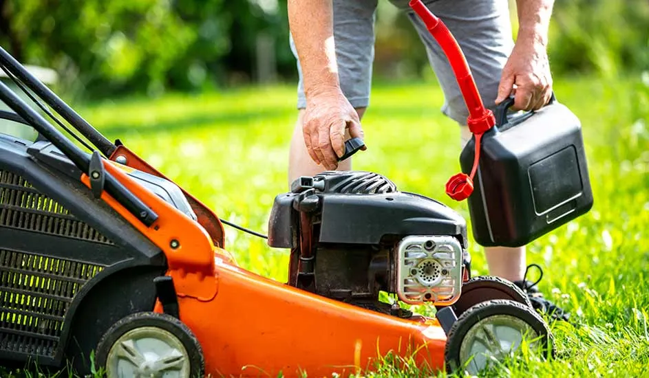 Een persoon vult een oranje grasmaaier bij met benzine in een groene tuin. Het merk van de grasmaaier is niet zichtbaar.