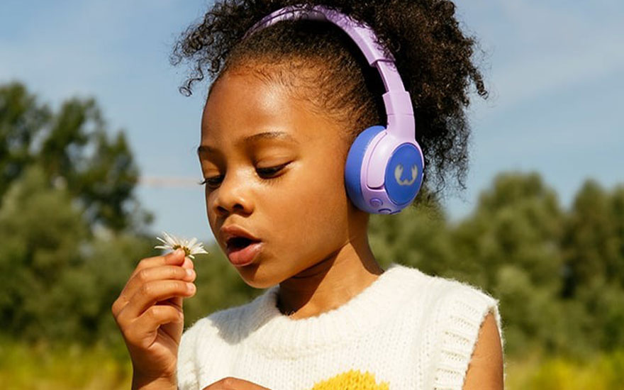 Meisje met paarse hoofdtelefoon ruikt aan witte bloem. Ze draagt een wit vest met een gele bloem. De achtergrond is blauwe lucht en groene bomen.