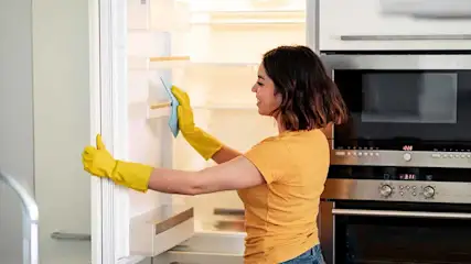 Een vrouw met gele handschoenen maakt een witte koelkast schoon met een blauwe doek in een keuken met apparatuur.