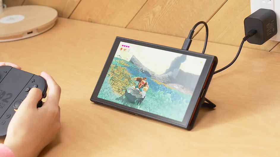 Een draagbare spelcomputer staat op een tafel, aangesloten op een zwarte stroomadapter in een stopcontact. Een persoon gebruikt een controller.