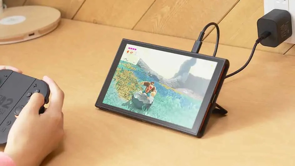 Een draagbare spelcomputer staat op een tafel, aangesloten op een zwarte stroomadapter in een stopcontact. Een persoon gebruikt een controller.