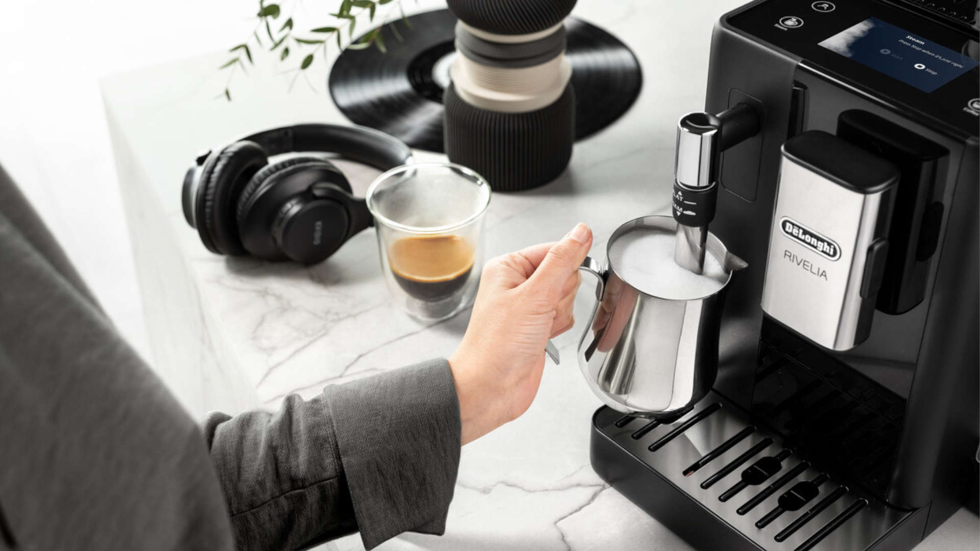 Op een marmeren aanrecht staat een Delonghi Rivelia koffiezetapparaat, een glas espresso, een melkkannetje, een koptelefoon en een speaker.