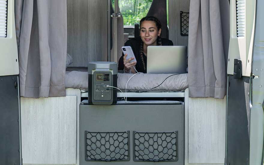 Een vrouw ligt ontspannen in een camper, werkt op haar laptop en laadt haar telefoon op via een draagbare stroomgenerator naast het bed.