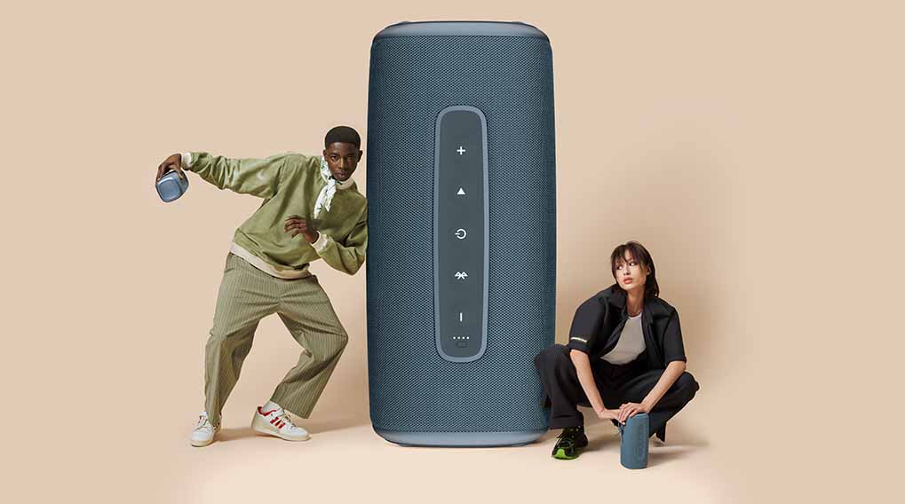 Een grijze luidspreker staat centraal tussen twee mensen op een lichtbeige achtergrond. Links staat iemand met een kleinere luidspreker.