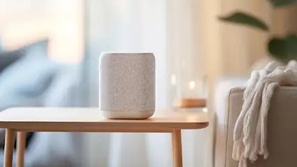 Een grijze speaker staat op een houten bijzettafel in een kamer met een bank en een deken.