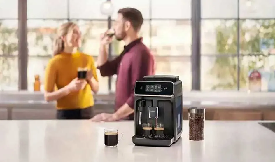 Een zwarte koffiemachine staat op een keukenblad met kopjes koffie en bonenpot, terwijl twee mensen op de achtergrond drinken.