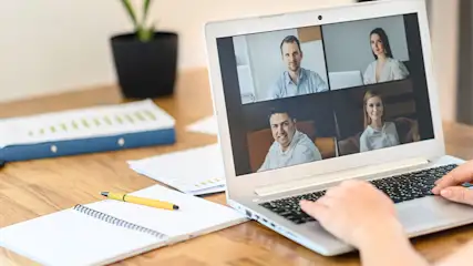 Laptop met videovergadering, vier deelnemers zichtbaar. Handen typen op toetsenbord, notitieboek, pen en plant op tafel.