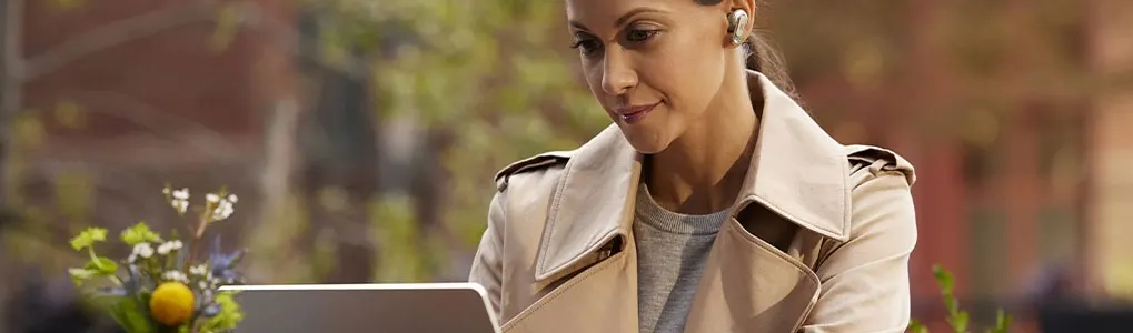 A person in a beige coat uses a laptop; a small flower arrangement sits beside the computer.