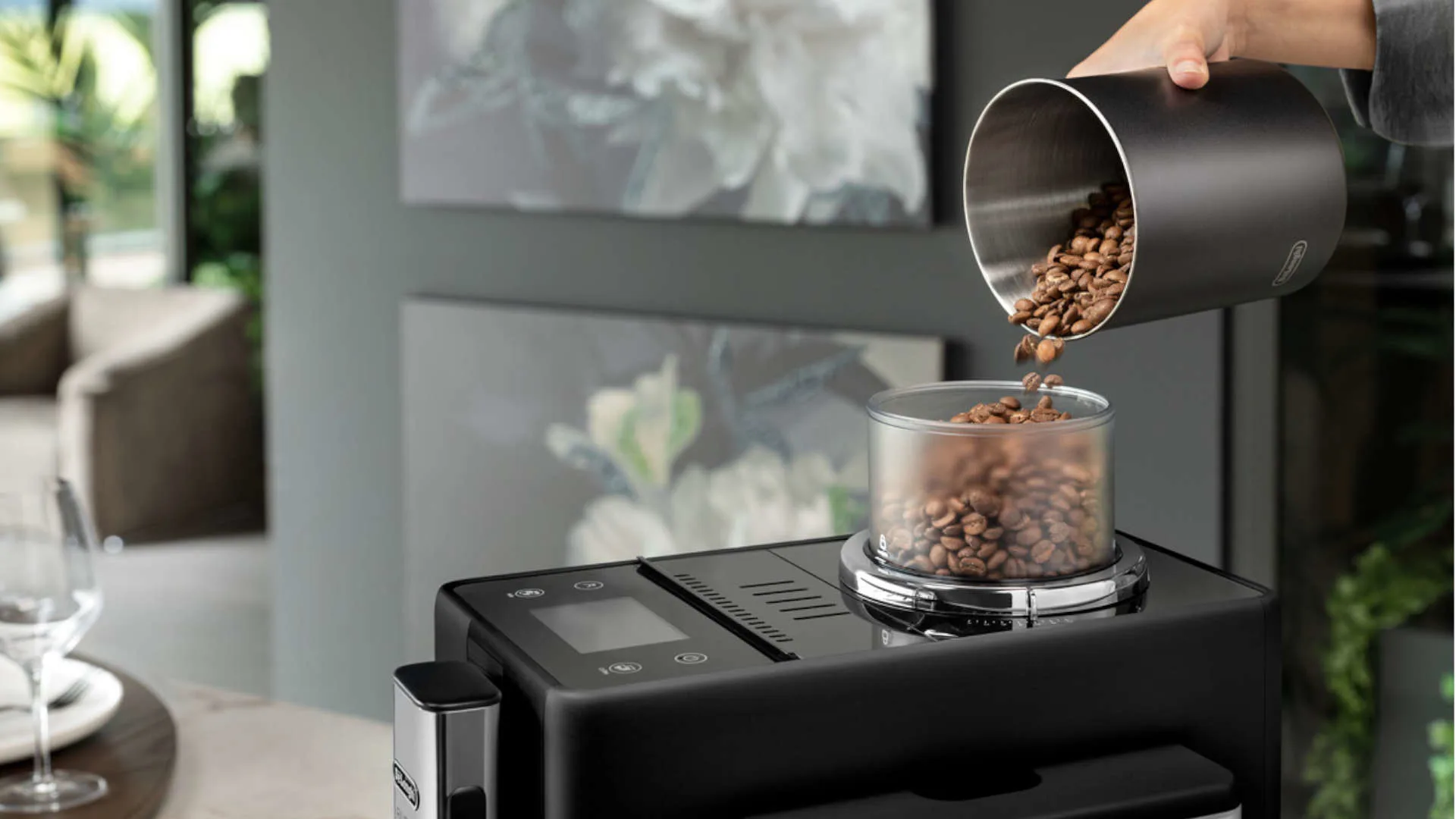 Een zwarte koffiemachine met een hand die koffiebonen in een transparant reservoir giet.