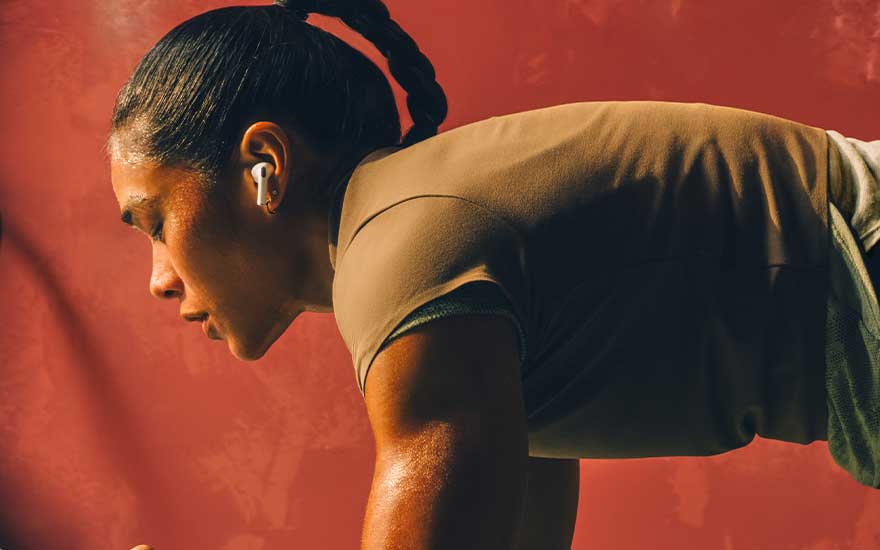 Athletic woman trains intensely against a red backdrop, sweaty and focused, wearing wireless earbuds while holding a demanding workout position.