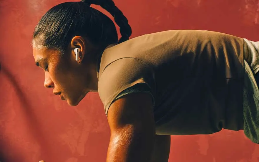 Athletic woman trains intensely against a red backdrop, sweaty and focused, wearing wireless earbuds while holding a demanding workout position.