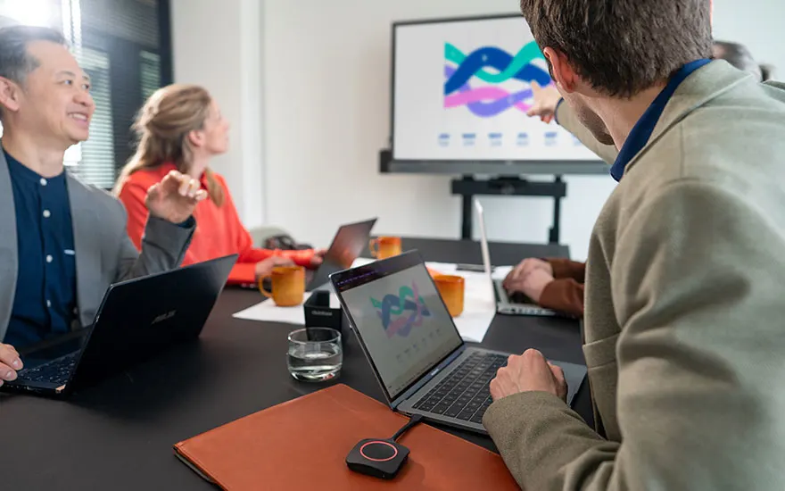 Moderne vergaderruimte met collega's rond tafel, ClickShare dongle bij laptop, groot scherm toont kleurrijke grafieken voor productieve meeting.