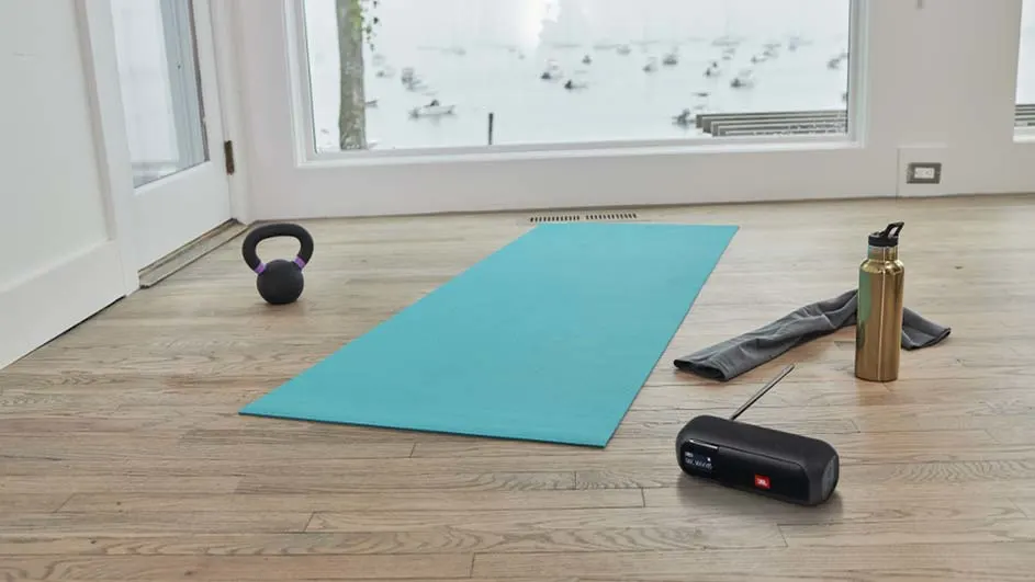 Een blauwe yogamat ligt op een houten vloer, met een kettlebell, waterfles, handdoek en speaker ernaast, voor een raam met sneeuw.